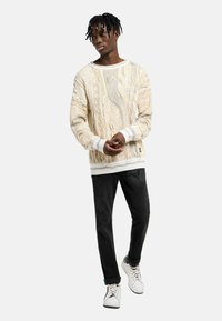 Strickpullover in Creme mit strukturiertem Muster, ausgestattet mit gerippten Bündchen und Saum. Kombiniert mit schwarzen Hosen und weißen Sneakern, stehende Pose.