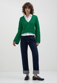 Maglione verde a collo a V con lavorazione a rotture, abbinato a jeans blu scuro con risvolti. Il modello indossa mocassini neri e una camicia bianca.