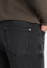 Jeans de denim negro con un acabado texturizado, que presentan un bolsillo trasero curvado con remaches metálicos y una vista cercana de la cinturilla.
