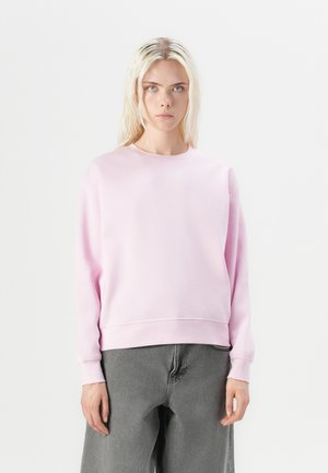 Pull en coton rose avec un col rond, des épaules tombantes et des poignets côtelés, associé à un pantalon gris foncé ample.