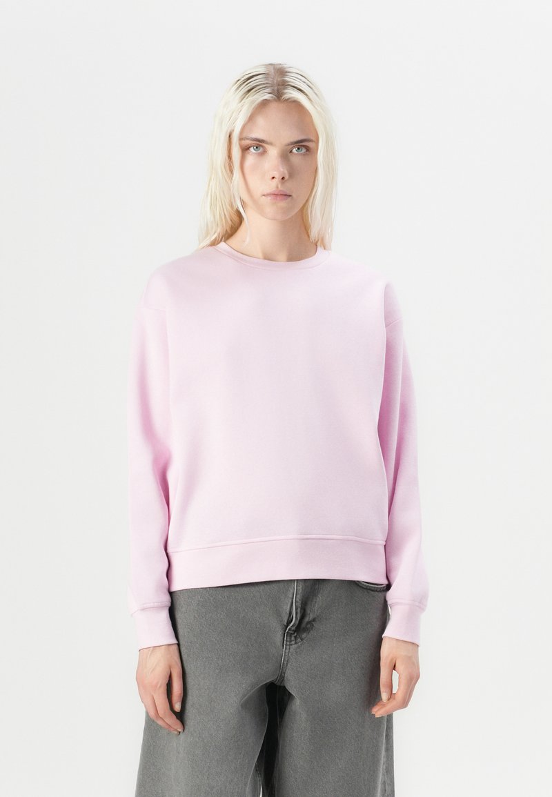 Felpa di cotone rosa con colletto rotondo, spalle cadenti e polsini a costine, abbinata a pantaloni grigi scuri morbidi.