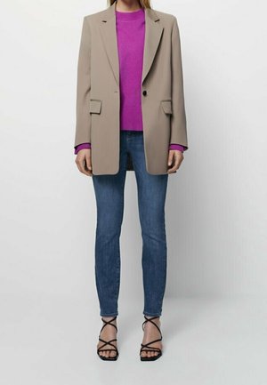 Personne portant un blazer beige, un pull violet vif, un jean skinny bleu et des sandales à talons noires avec lanières, posant devant un fond uni.