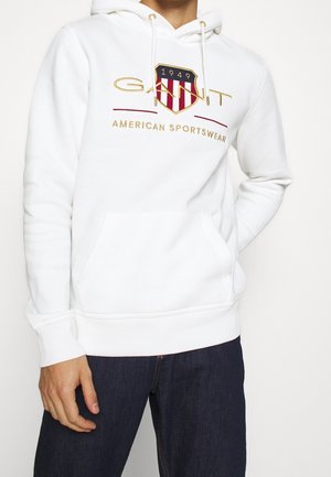 Sudadera blanca con un bolsillo frontal tipo canguro, capucha con cordón y un logo bordado con acentos en rojo y amarillo. Tejido suave, diseño casual.