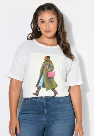 Femme aux cheveux bruns ondulés portant des boucles d'oreilles créoles, un T-shirt blanc avec un visuel d'une personne en manteau vert et sac rose, et un jean bleu.