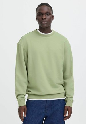 Mann trägt hellgrünes Sweatshirt mit Rundhalsausschnitt über weißem Hemd und blauer Jeans, steht vor einem weißen Hintergrund.