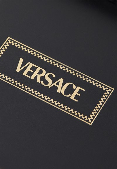 Superficie texturizada negra con una etiqueta rectangular bordeada en dorado que muestra "VERSACE" en una fuente en negrita, con un acento estampado a lo largo de los bordes.