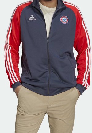 Mann trägt eine marineblaue und rote Adidas-Jacke mit dem FC Bayern München-Logo, beige Hose und hat die Hände in den Taschen.