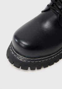 Bota de couro preta com sola grossa e com ranhuras destacadas e detalhe de costura branca, close-up na ponta e área dos atacadores contra fundo branco.