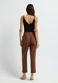 Vrouw staand met de rug naar de kijker, gekleed in een zwarte mouwloze top, bruine op maat gemaakte broek en beige kitten heels tegen een effen achtergrond.