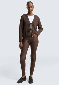 Brun stickad kofta med knytning framtill, bärs över en vit topp, kombinerad med bruna figursydda byxor och svarta loafers. Mjuk textur.