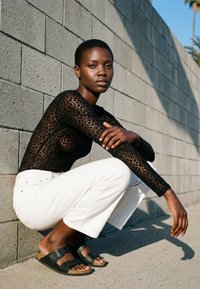 Vrouw met kort haar, die een doorschijnende zwarte top met luipaardprint, witte broek en zwarte sandalen draagt, hurkt naast een betonnen blokmuur in het zonlicht.