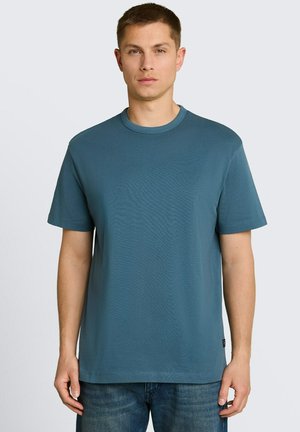 Młody mężczyzna ubrany w jednokolorowy turkusowy t-shirt z krótkim rękawem i niebieskie dżinsy, stojący na tle jasnoszarego tła.