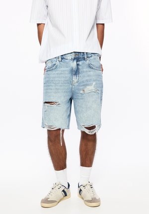 Person trägt hellblaue, abgenutzte Denim-Shorts, weiße Socken und beige Sneaker mit blauen Akzenten, steht mit den Händen am Rücken.