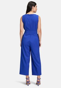 Kráľovsky modrý overal s bezrukávovým dizajnom, širokými nohavicami, opasok v páse a zapínanie na zips na chrbte. Má hladkú, ľahkú tkaninu.