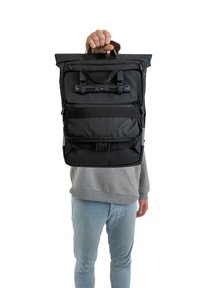 Svart rulltoppsryggsäck med flera remmar, slät yta, sidoficka med dragkedja och hållbara handtag, med en minimalistisk design.