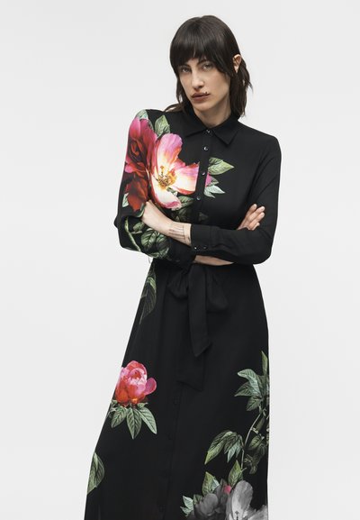 Mujer de cabello oscuro con un vestido negro de manga larga con grandes estampados florales en rosa, rojo y blanco, brazos cruzados, expresión neutra.