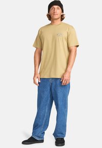 Beige katoenen t-shirt met korte mouwen, voorzien van een klein logo op de borst; gecombineerd met loszittende blauwe spijkerbroek en zwarte schoenen.