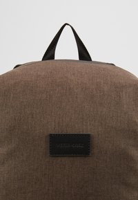 Brun, strukturerad tygryggsäck med svart handtag upptill och svart rektangulär märkesetikett centrerad på framsidan.