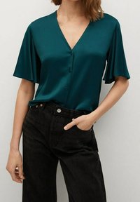 Blusa di raso teal con scollatura a V, maniche corte a sbuffo e dettagli con bottoni, abbinata a jeans neri a vita alta. Tessuto liscio.