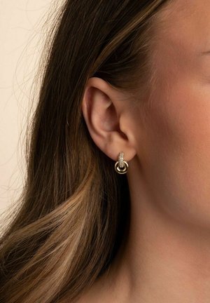 Goldfarbene Ohrringe mit einem kreisförmigen Design, das einen strukturierten Ring und glatte Akzente aufweist. Tragen von einem Modell am Ohr, zusammen mit lockigem Haar.