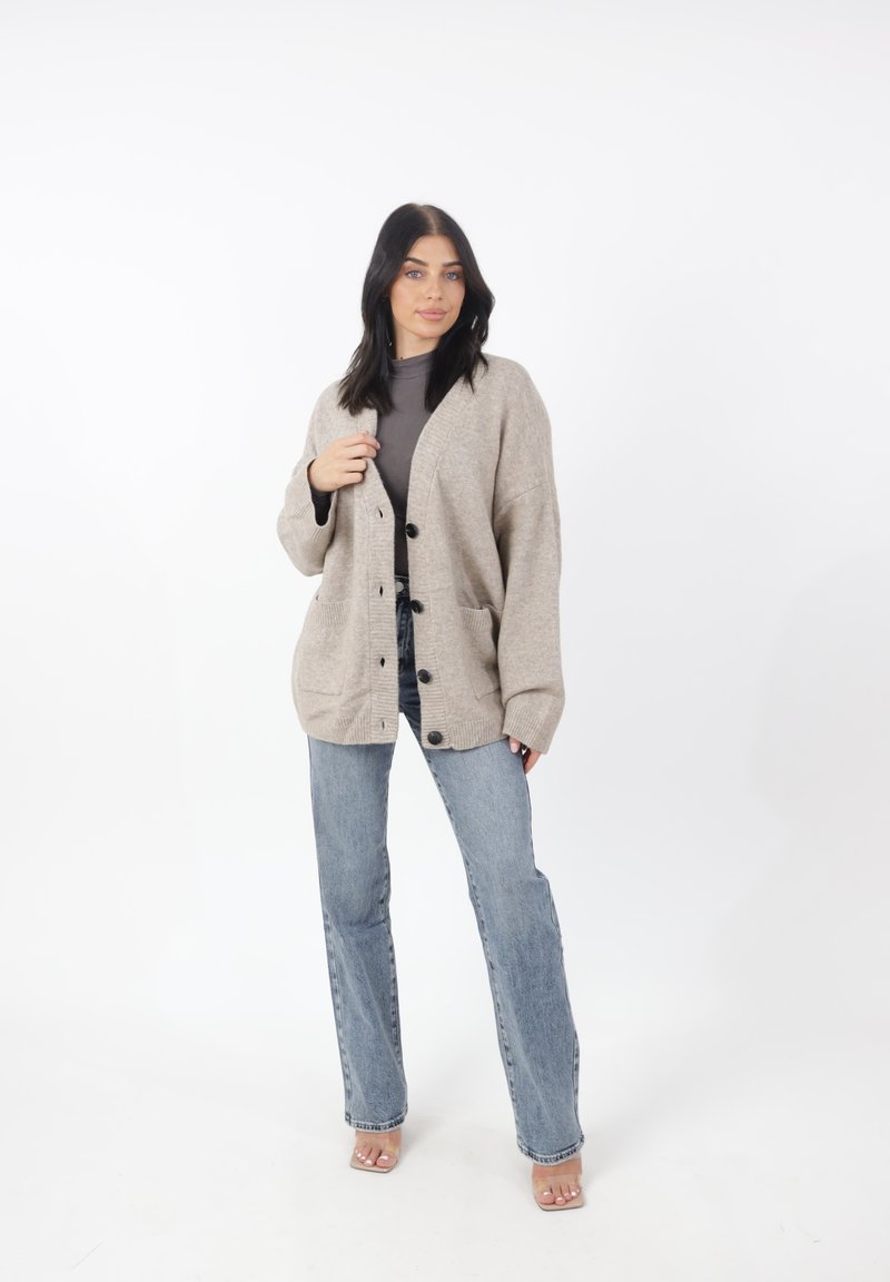 Cárdigan beige de punto con grandes botones negros, mangas anchas y bolsillos laterales, usado sobre una blusa gris y combinado con jeans de un azul claro.