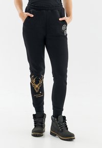 Čierne tepláky s elastickým pásom, zúžený strih, s grafickým motívom zlatého jelena a textom "EXPECTO PATRONUM" na ľavej nohe. V kombinácii s čiernymi čižmami.