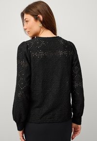Blouse en dentelle noire avec un motif texturé, longues manches bouffantes et col rond. Présente des découpes complexes et une silhouette ajustée.