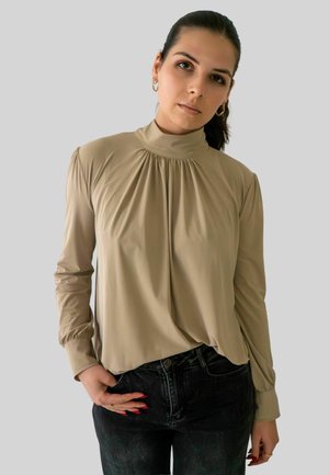 Femme aux cheveux bruns attachés, portant un haut beige à col montant et un jean noir, se tenant avec une main dans la poche devant un fond uni.