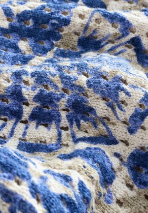Knit throw blanket featuring intricate blue floral patterns on a cream background. Textured yarn with noticeable stitching and lightweight design.