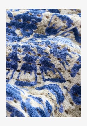 Strickwurfdecke mit komplexen blauen Blumenmustern auf einem cremefarbenen Hintergrund. Strukturierter Garn mit auffälligen Nähten und leichtem Design.