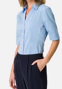 Hellblaues kurzärmeliges Hemd mit Button-Down-Kragen und Frontleiste; kombiniert mit dunklen Hosen, die eine glatte Textur und eine maßgeschneiderte Passform zeigen.