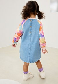 Spijkerjurk met roze, patroonmouwen met kleurrijke cartoonfiguren. Achterknopen en een afgeronde zoom maken het ontwerp compleet.