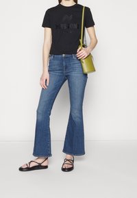 Musta puuvillainen t-paita, jossa "MISSONI"-logo, siniset levenevät farkut, mustat nauhalla kiinnitettävät sandaalit ja pieni vihreä käsilaukku olkahihnalla.