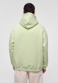 Lichtgroene oversized hoodie met een trekkoord in de kap, ribgebreide manchetten en zoom. Zachte textuur en casual pasvorm. Achteraan zijn de naaddetails zichtbaar.