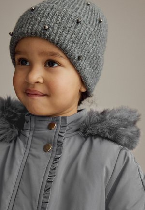 Bonnet en tricot gris avec des accents métalliques et une texture côtelée, associé à un manteau gris avec un devant froncé et une capuche bordée de fourrure.