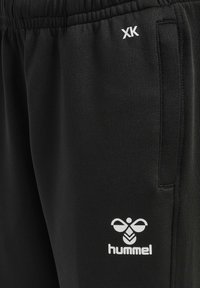 Zwarte sportbroek van gladde stof, met een elastische tailleband, zijzakken en een wit 'Hummel' logo nabij de onderkant.