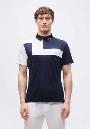 Ung mann iført en marineblå polo med hvitt geometrisk mønster og hvite bukser, stående vendt fremover foran en ensfarget bakgrunn.