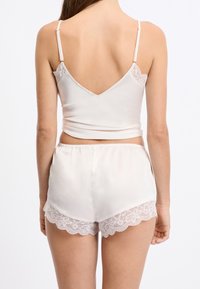 Dwuczęściowy zestaw z jedwabiu w bladoróżowym kolorze, składający się z topu na cienkich ramiączkach z dekoltem w kształcie litery V na plecach oraz szortów z falbanką z koronki na dole i regulowanymi ramiączkami.