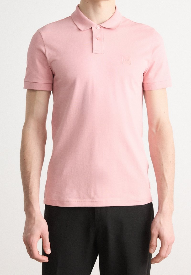 Homme portant un polo rose clair à manches courtes avec deux boutons et un pantalon noir, visible du cou aux hanches.