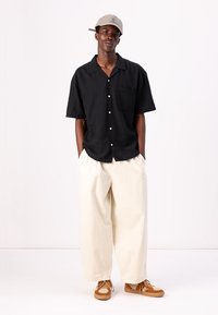 Camisa negra de manga corta con un bolsillo frontal, combinada con pantalones beige holgados y zapatos marrones, complementada por una gorra gris.
