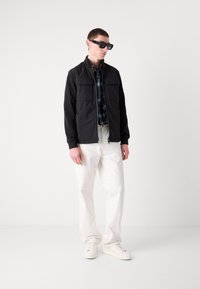 Jachetă ușoară neagră cu fermoar, cu două buzunare pe piept, purtată peste o cămașă cu carouri albastre și cu pantaloni crema și adidași.