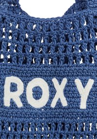Borsa blu lavorata a uncinetto realizzata con fibre di cotone, caratterizzata da una grande scritta bianca "ROXY", con trama aperta e parte superiore arrotondata.