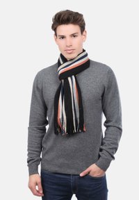 Jeune homme portant un pull gris, un jean noir et une écharpe rayée noire avec des accents bleus, blancs et orange, posant avec une main dans la poche.