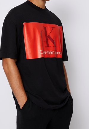 Mężczyzna noszący czarną koszulkę z krótkim rękawem, z dużym czerwonym prostokątnym logo na piersi z napisem „cK” i „Calvin Klein Jeans”.