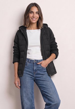 Femme souriante portant une veste matelassée noire sur une chemise blanche et un jean bleu, debout avec une main dans la poche devant un fond uni.