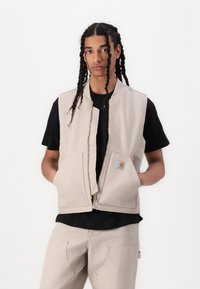 Beige canvas vest med glidelås foran, to store frontlommer og et logo-lapp. Båret over en svart skjorte, sammen med beige bukser.