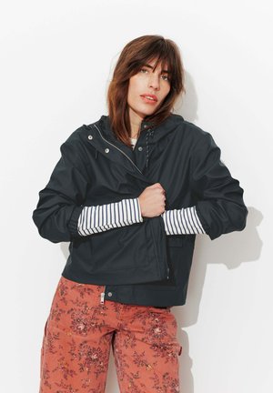 Femme portant une veste à capuche noire par-dessus un haut à manches longues rayé et un pantalon floral orange rouille, debout devant un fond blanc.