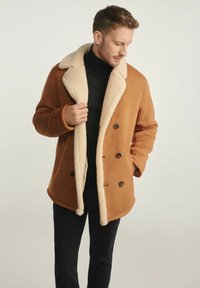 Brązowy płaszcz dwurzędowy z kremowym kołnierzem z baranka, teksturowana tkanina i brązowe guziki, noszony na czarnym golfie.