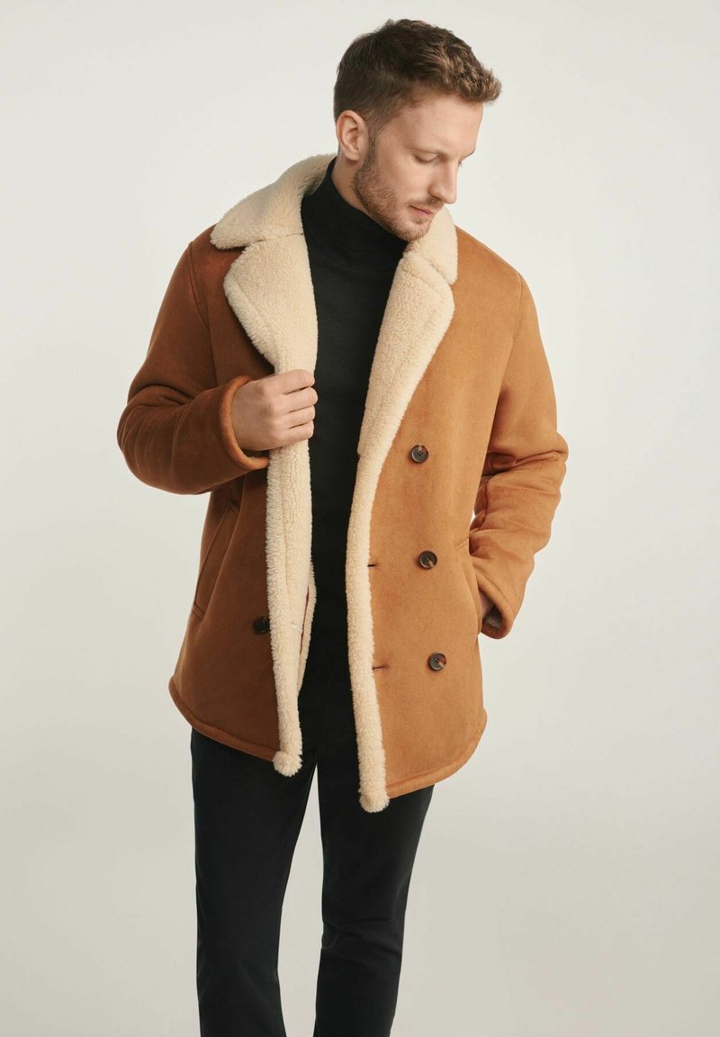 Brązowy płaszcz dwurzędowy z kremowym kołnierzem z baranka, teksturowana tkanina i brązowe guziki, noszony na czarnym golfie.