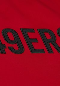 Roter Stoff mit großen schwarzen gestickten Buchstaben, die "49ERS" in einer fetten, klaren Schrift anzeigen. Die Stickerei ist auffällig und detailliert.